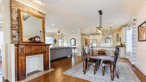 Un vaste espace dédié à la salle à manger, avec une table et des chaises en bois, une cheminée surmontée d’un miroir, et une cuisine moderne en arrière-plan.