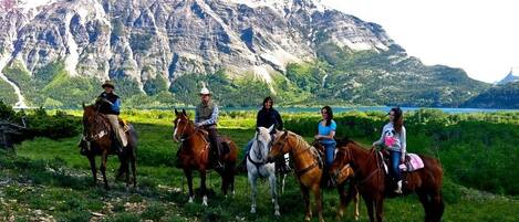 A group of people on horseback in a grassy field with a mountainous backdrop.