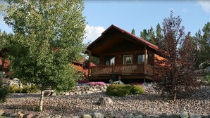 A wooden cabin with a balcony, surrounded by a garden with various plants and rocks. - #4 - FISHERMAN CABIN – BEAUTIFUL PRIVATE SETTING, 6 MILES TO GLACIER PARK (Columbia Falls)