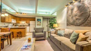 Un vaste espace de vie comprenant un canapé, une table basse et une table à manger avec des chaises. On y trouve également un coin cuisine avec des meubles en bois et un four à micro-ondes. Un élément décoratif mural et une lampe sont également visibles.