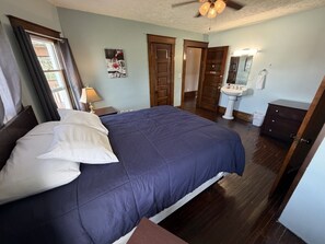 A bedroom with a large bed, a nightstand, a ceiling fan, a window with curtains, and a door leading to another room.