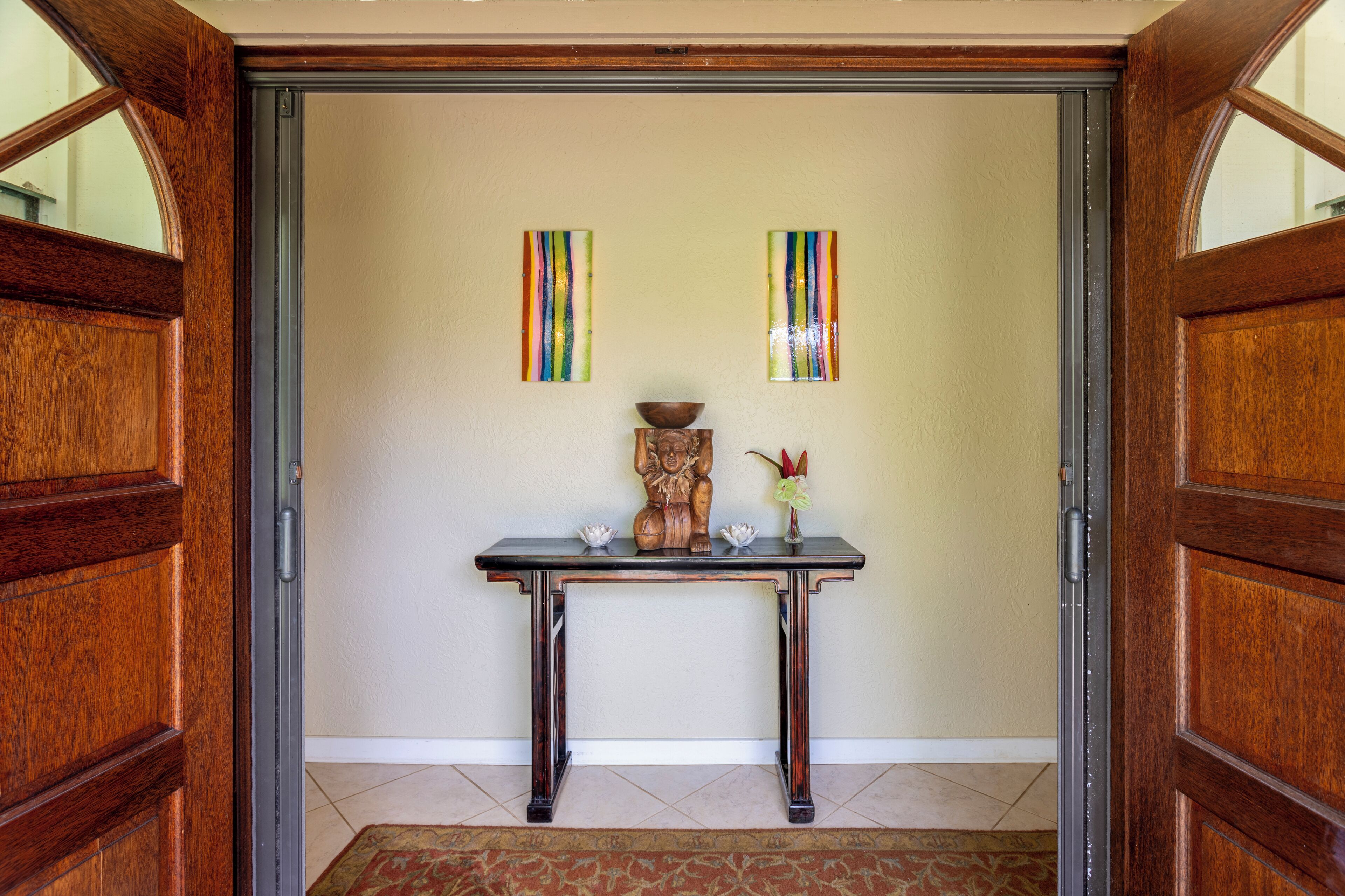 Une porte en bois avec un panneau de verre, une console en bois ornée d’un vase et de fleurs, et deux éléments de décoration murale à rayures verticales.