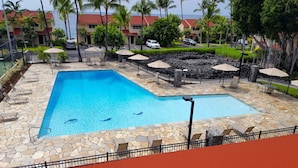 Una piscina con baldosas de piedra, rodeada de sillones de descanso y sombrillas, con vista a un edificio de techo rojo y palmeras al fondo.