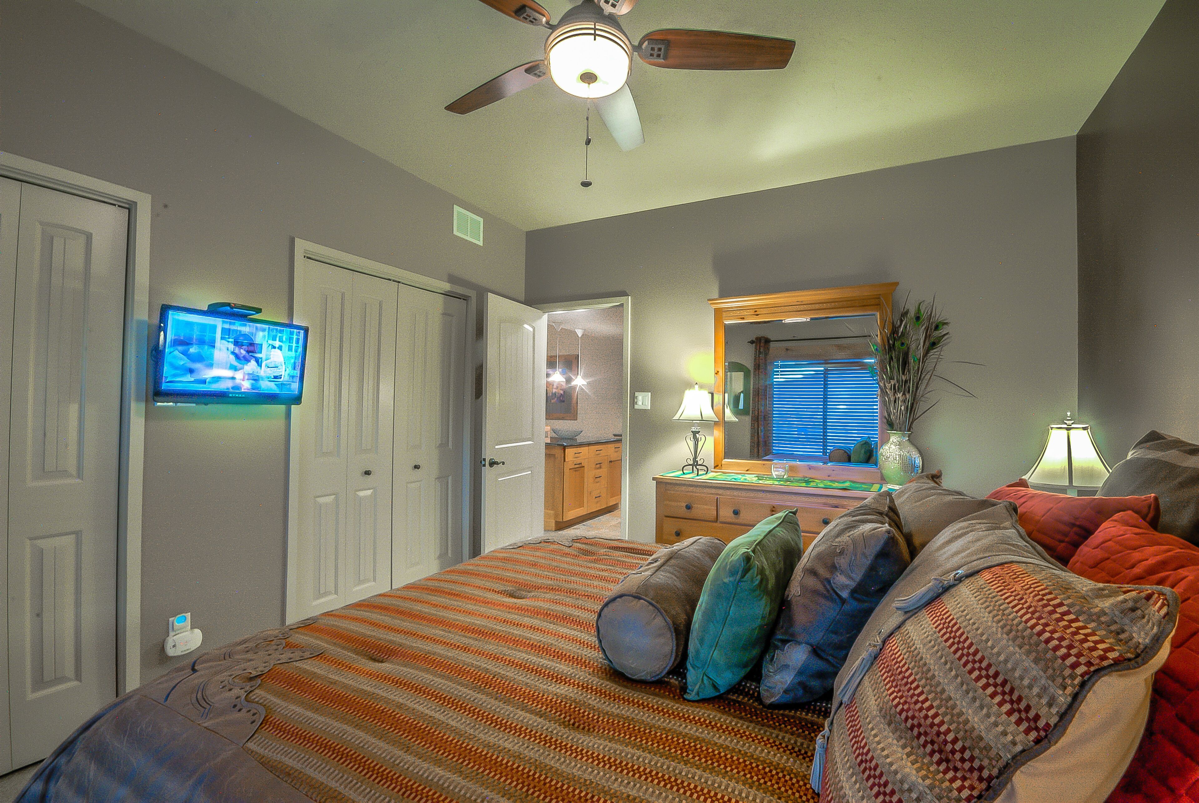 A bedroom with a bed, pillows, a ceiling fan, a TV, a dresser, a mirror, and a lamp.