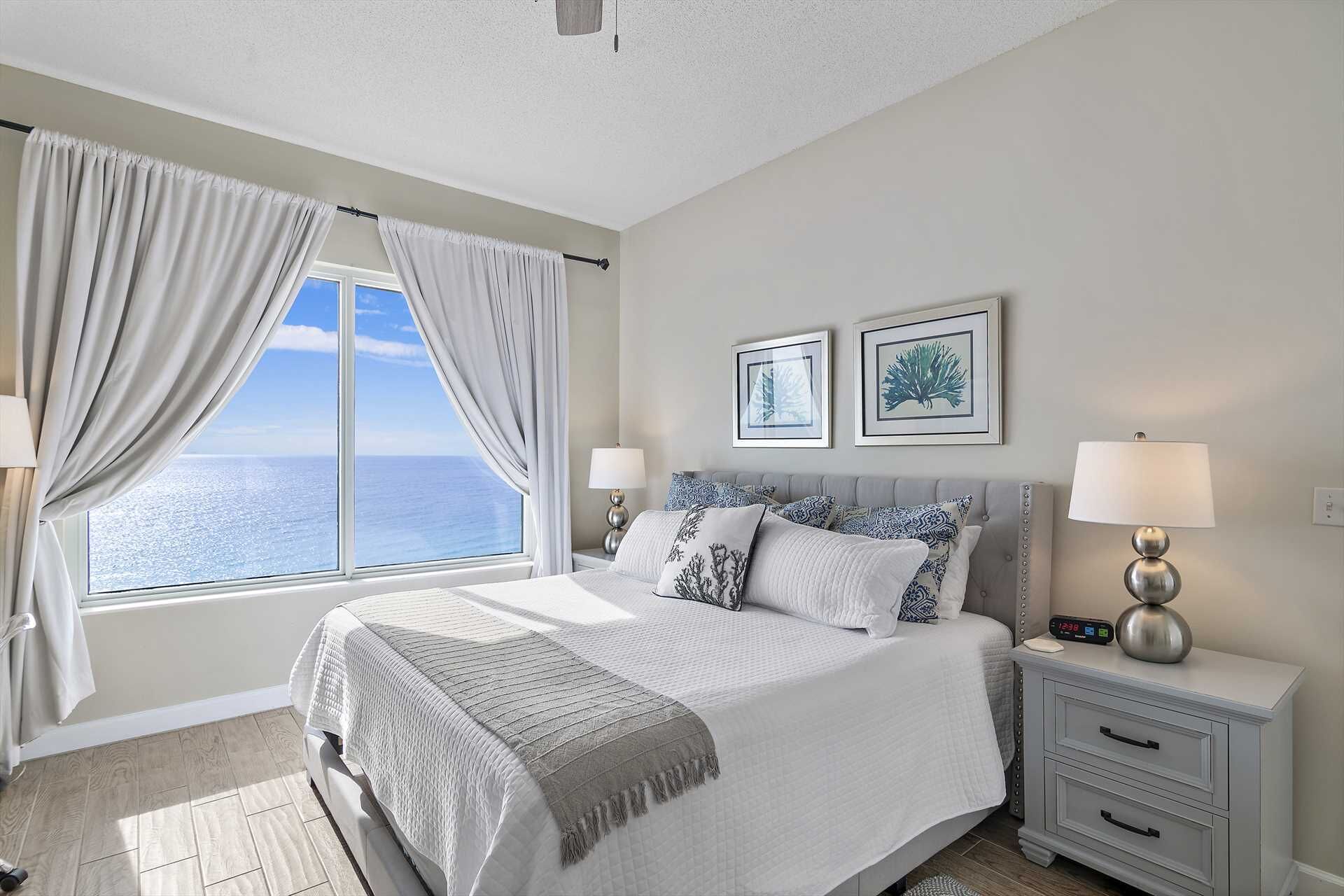 A bedroom with a large bed, two bedside tables with lamps, a view of the sea through a window, and two framed pictures on the wall.