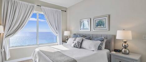 A bedroom with a large bed, two bedside tables with lamps, a view of the sea through a window, and two framed pictures on the wall.