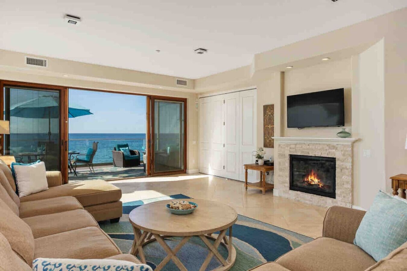 A spacious living room with a fireplace, a television mounted above it, and a large sliding glass door leading to a balcony with a view of the sea.