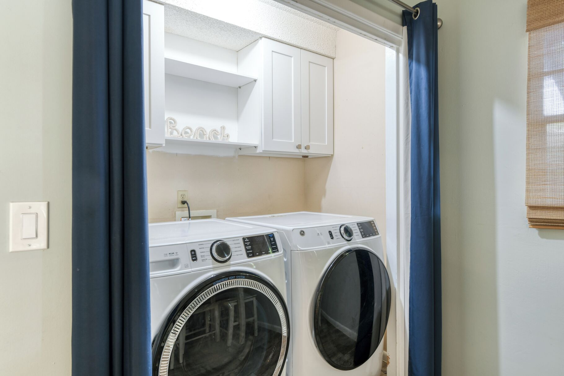 A laundry room with two front-loading washing machines and a dryer, white cabinets above, and a power outlet on the wall.