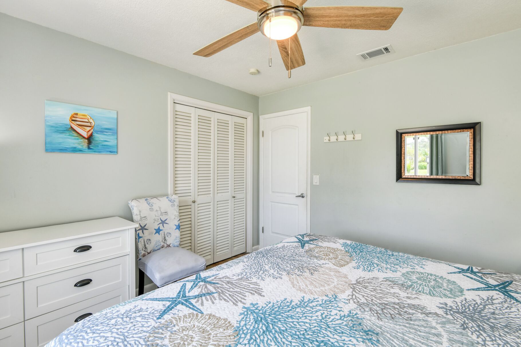 A bedroom with a bed, a ceiling fan, a door, a dresser, a painting, and a mirror.