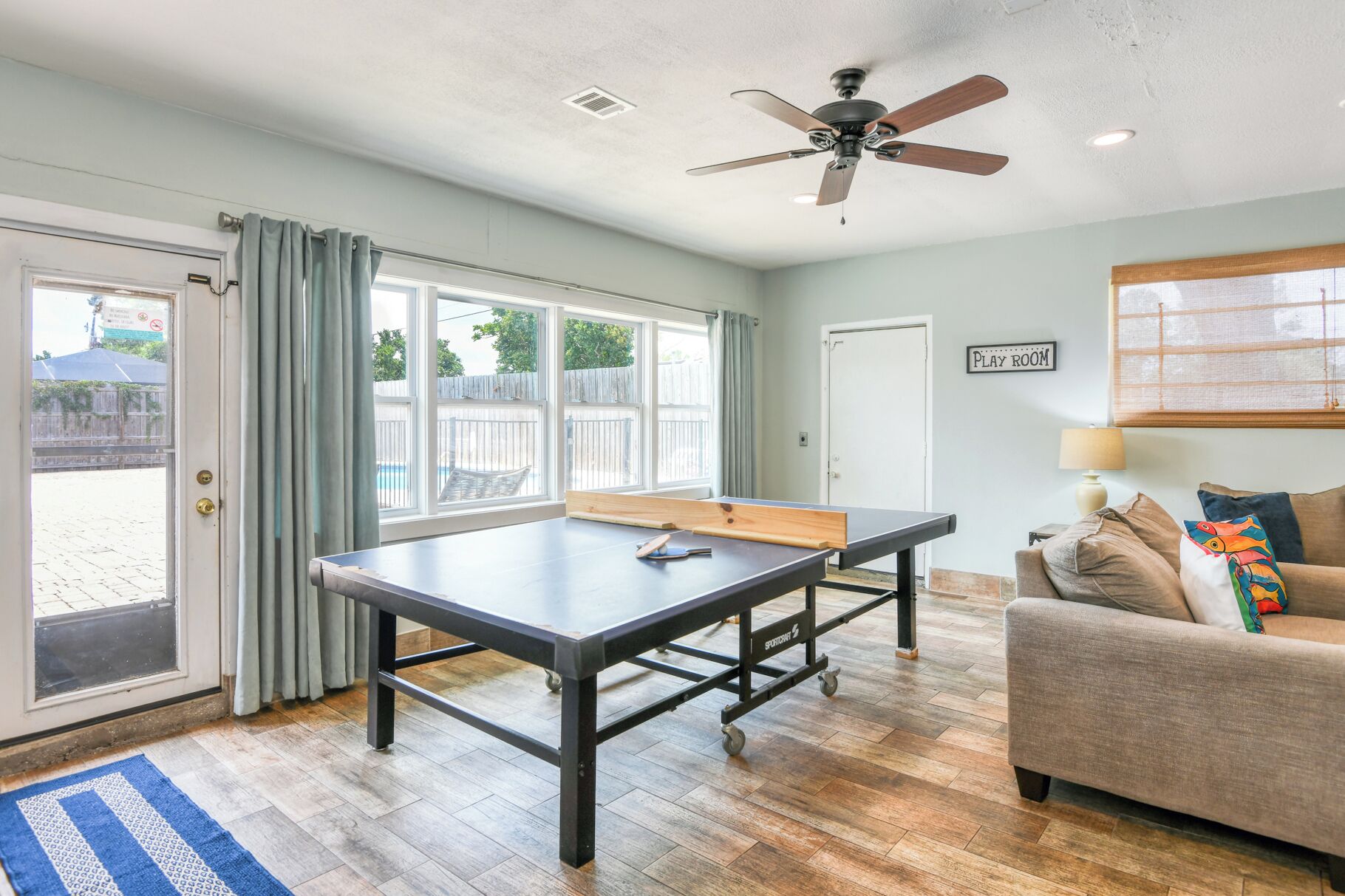 A room with a ping pong table, a ceiling fan, a couch, and a door leading to a patio.