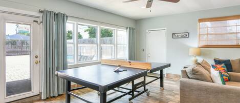A room with a ping pong table, a ceiling fan, a couch, and a door leading to a patio.