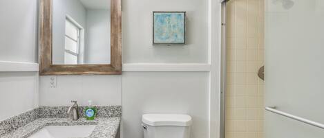 Un baño con lavamanos de granito, inodoro blanco, espejo enmarco y zona de ducha.