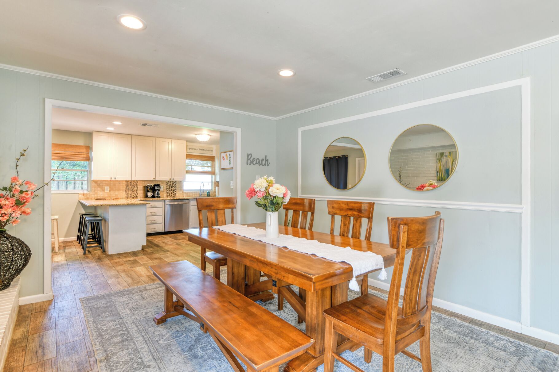A dining area with a wooden table and chairs, a kitchen in the background, and a wall with two round mirrors.