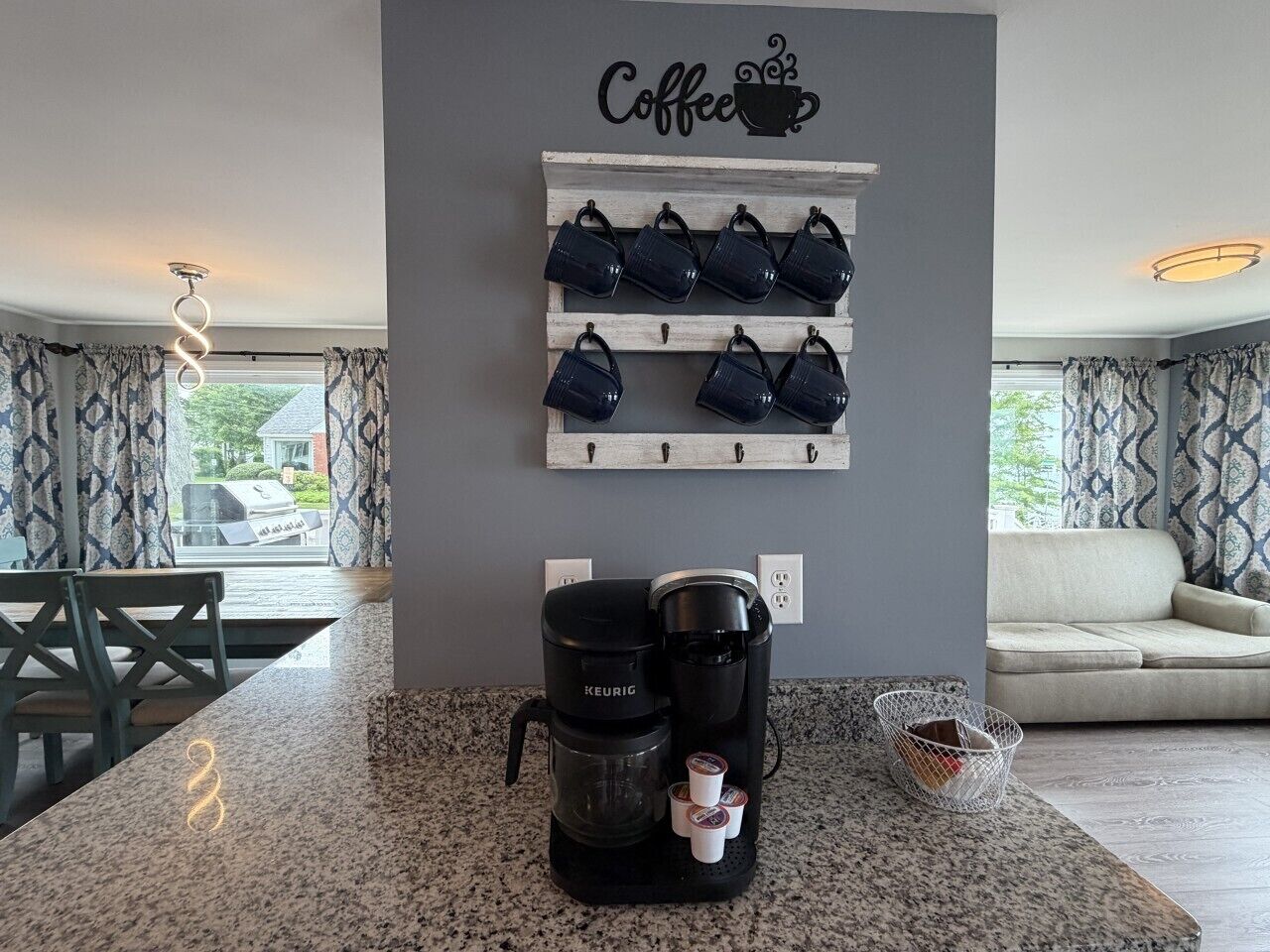 A kitchen with a coffee maker, a wall-mounted mug rack, and a dining area with a table and chairs.