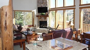 Un salon chaleureux avec une cheminée en pierre, des meubles en bois et de grandes fenêtres donnant sur l’extérieur.