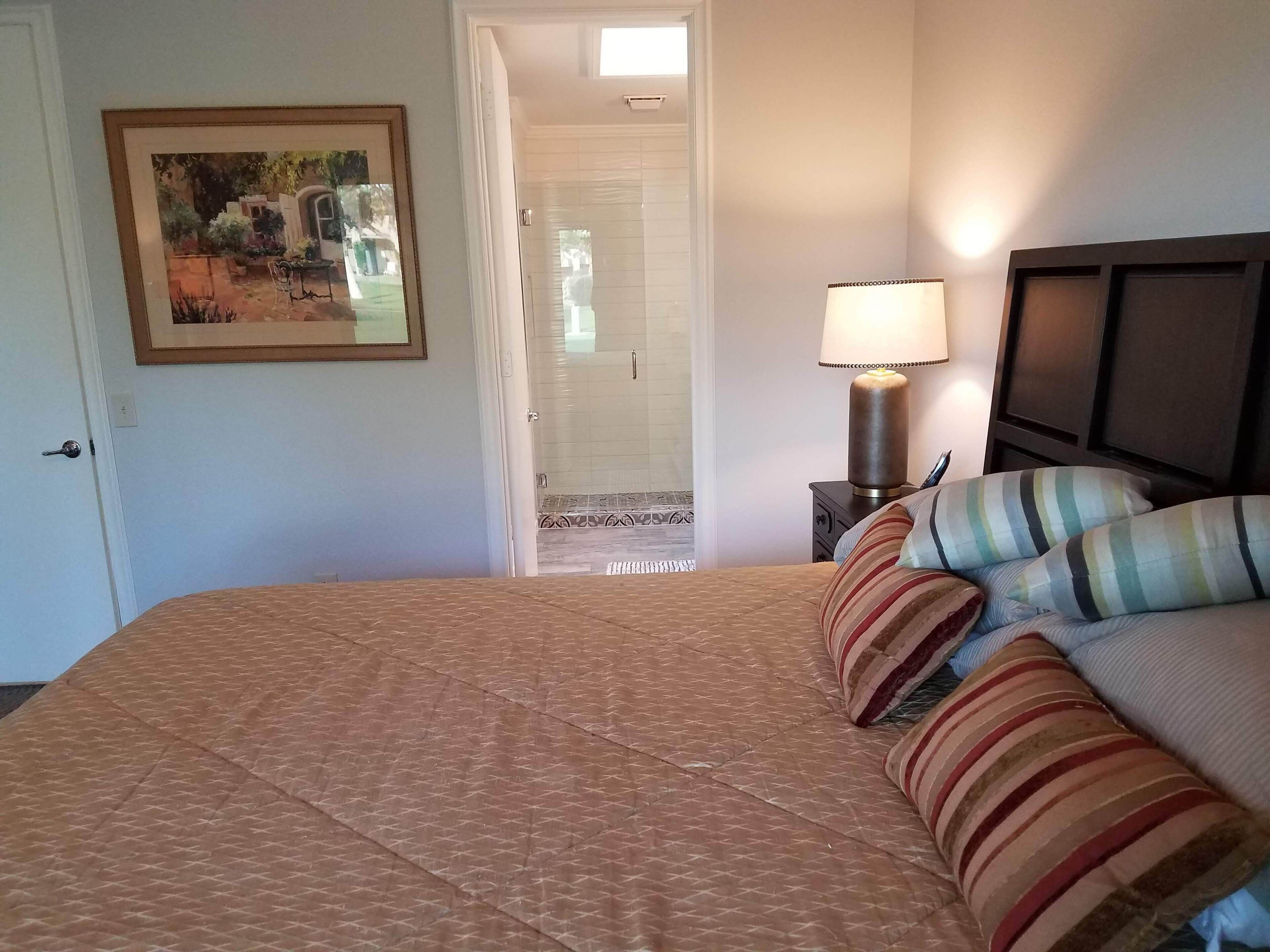 A hotel room with a bed, a nightstand with a lamp, a painting on the wall, and a door leading to a bathroom.