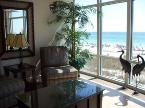 A living room with a view of the beach, a chair, a table, a lamp, and two decorative birds.