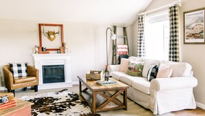 A cozy living room with a fireplace, a white sofa, a wooden coffee table, and a cowhide rug. - The Red Barn-Perfect GNP Base Camp. 5 Star Luxury on the farm. Clean & Comfy. (Columbia Falls)