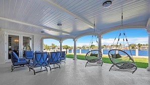 A covered outdoor seating area with blue chairs and a swing chair, overlooking a body of water and a cityscape.