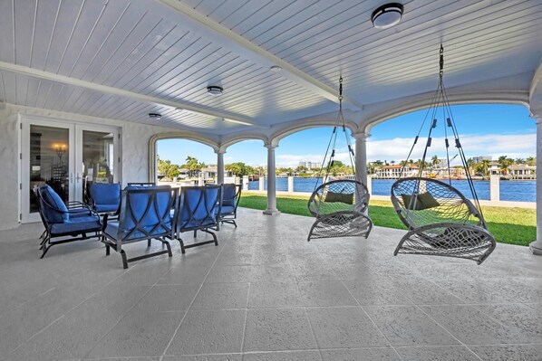 A covered outdoor seating area with blue chairs and a swing chair, overlooking a body of water and a cityscape. - Waterfront Estate with Pool and Dock (Fort Lauderdale)