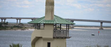A small tower structure with a green roof and a balcony, overlooking a body of water with a bridge in the background.