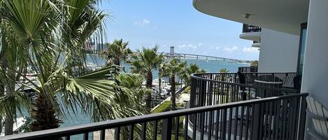 A balcony with a view of the sea, palm trees, and a bridge in the distance.
