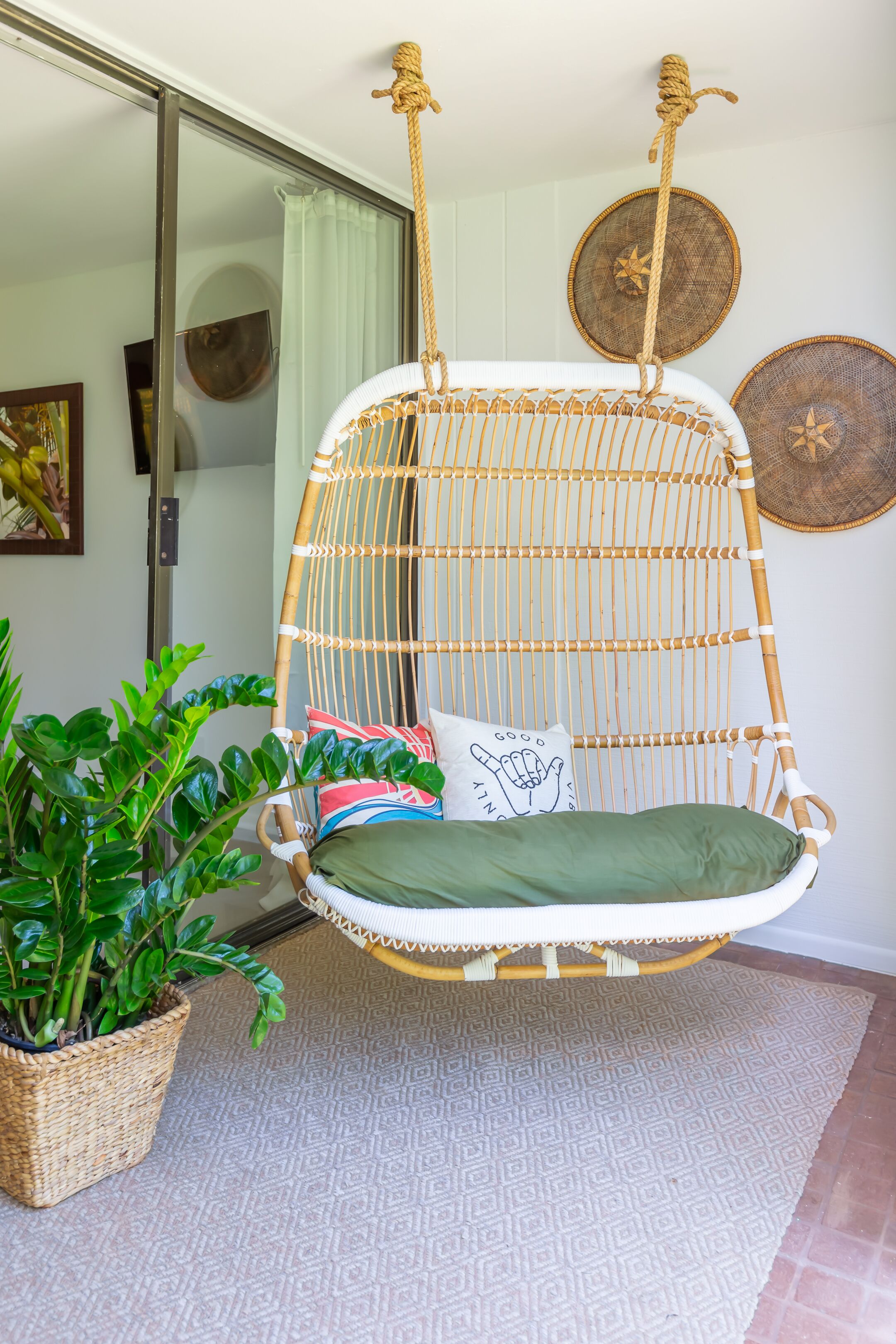 Una silla colgante de mimbre con un cojín verde, una planta en maceta y platos decorativos en la pared.