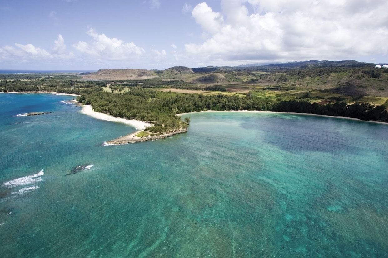 An aerial view of a coastal landscape with clear turquoise waters, a sandy beach, and a small island with dense vegetation.