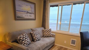 Una sala de estar con un sofá, cojines estampados, una mesa y un ventanal con vista a la playa y al océano.