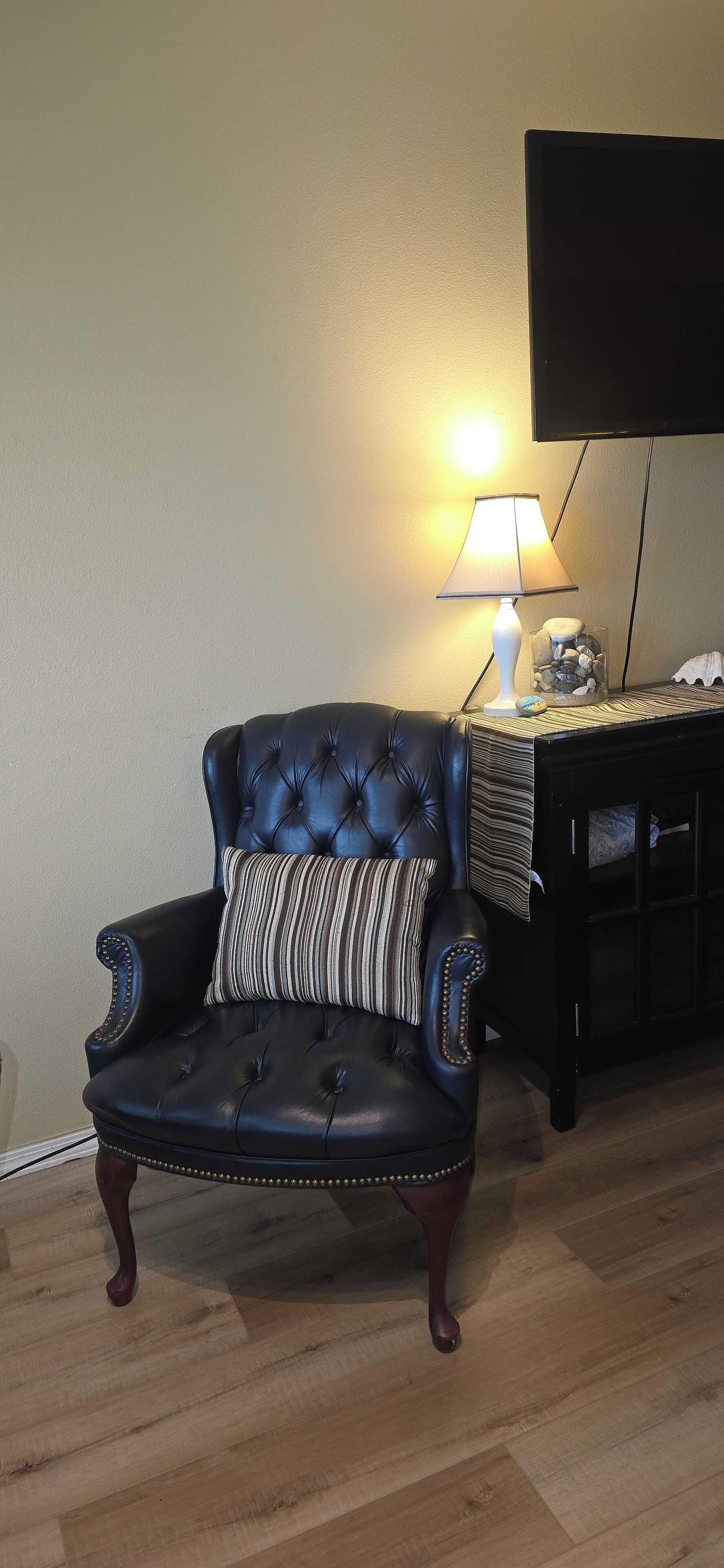A black leather armchair with a striped pillow, a lamp, and a glass container with stones on a wooden side table.