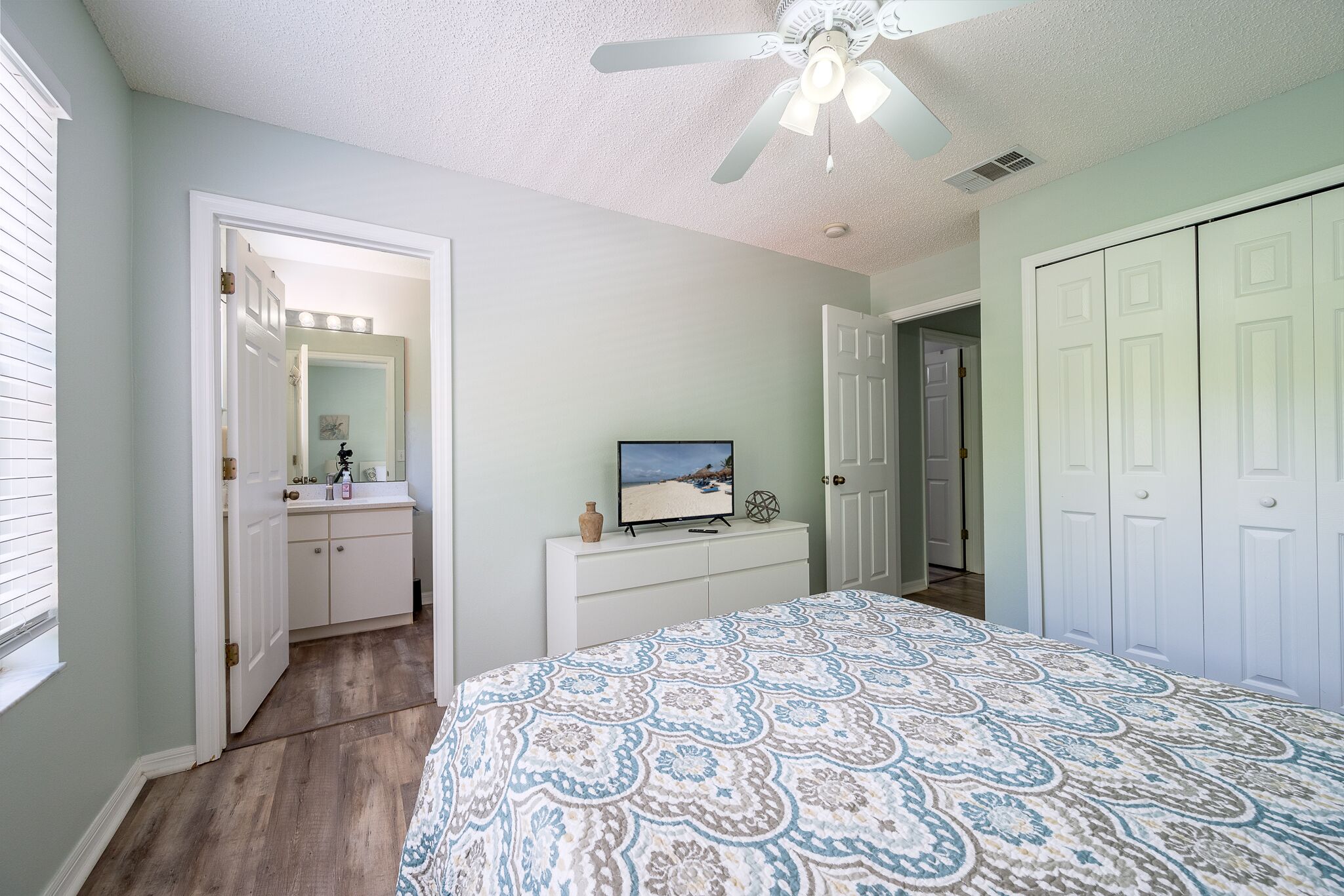 Une chambre à coucher comprenant un lit, une coiffeuse avec un miroir, un ventilateur de plafond et un placard.