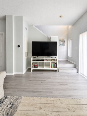 A modern living room with a flat-screen TV on a white entertainment unit, a staircase leading to an upper level, and a large window on the right. - 3 Bedroom Townhome in Gated Community. Pet Friendly! (Osceola County)
