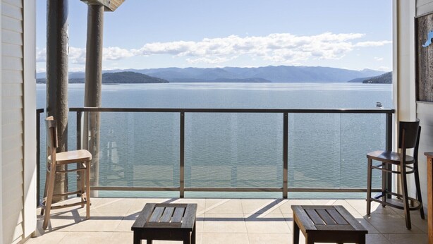 A balcony with a view of a lake and mountains, wooden furniture, and a metal railing. - Walk to Downtown, Top Floor- Seasons at Sandpoint (Sandpoint)