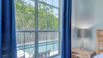 Una habitación con un amplio ventanal con vistas a una piscina, una mesita de noche de madera con una lámpara y una cama con colcha de diseño azul y blanco.
