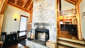A stone fireplace with a wooden mantel, a dining area with a high wooden ceiling, and a kitchen visible in the background.