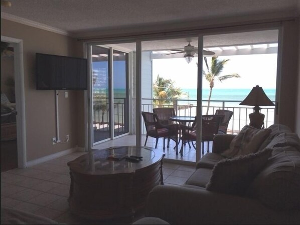 A living room with a sofa, a coffee table, and a TV on the wall. There is a balcony with a table and chairs, and a view of the ocean. - Direct Ocean View from Your Condo-No Contact Check-In (Key West)