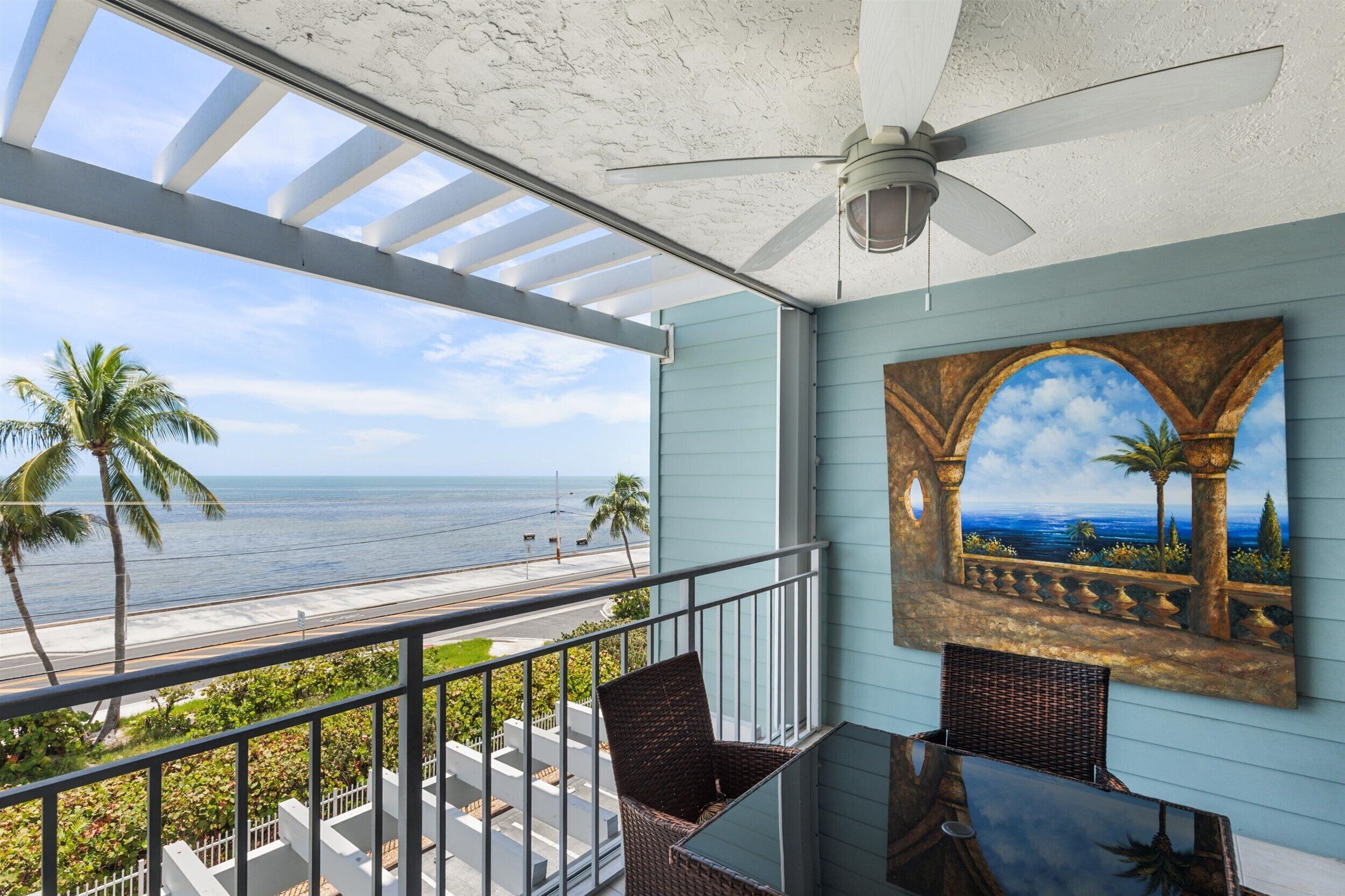 Un balcon avec vue sur l’océan, un ventilateur de plafond et un tableau représentant un paysage côtier.