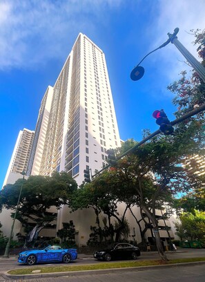 Un grand immeuble à l’architecture moderne, une rue bordée de voitures et des arbres au premier plan.