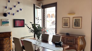 Ein Essbereich mit Holztisch, Stühlen und einer Blumenvase. Es gibt einen Holzschrank und einen Blick ins Freie durch ein Fenster.