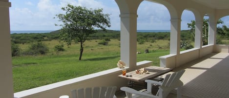 A covered outdoor seating area with white chairs and a table, overlooking a grassy landscape with trees and hills.