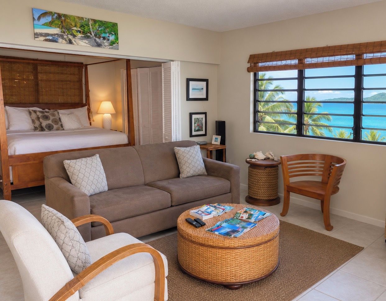 A room with a bed, a sofa, a round wicker ottoman, and a window with a view of the ocean.