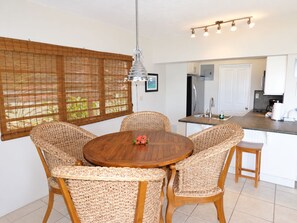 Un coin repas avec une table ronde en bois entourée de chaises en osier, un luminaire suspendu et une vue sur la cuisine à travers une fenêtre dotée de stores en bambou.