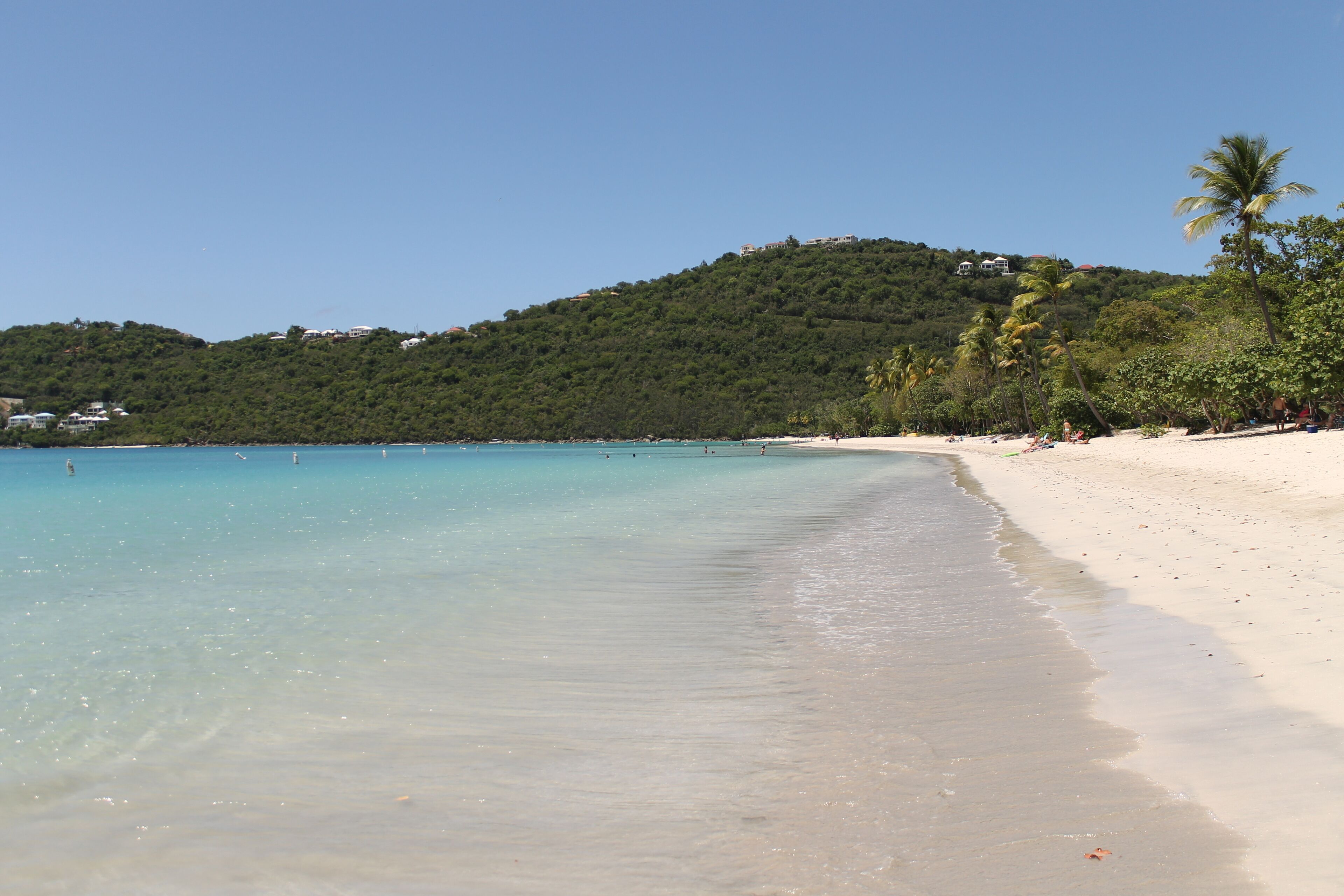 A beach with clear turquoise water, white sand, and a hill with buildings in the background.