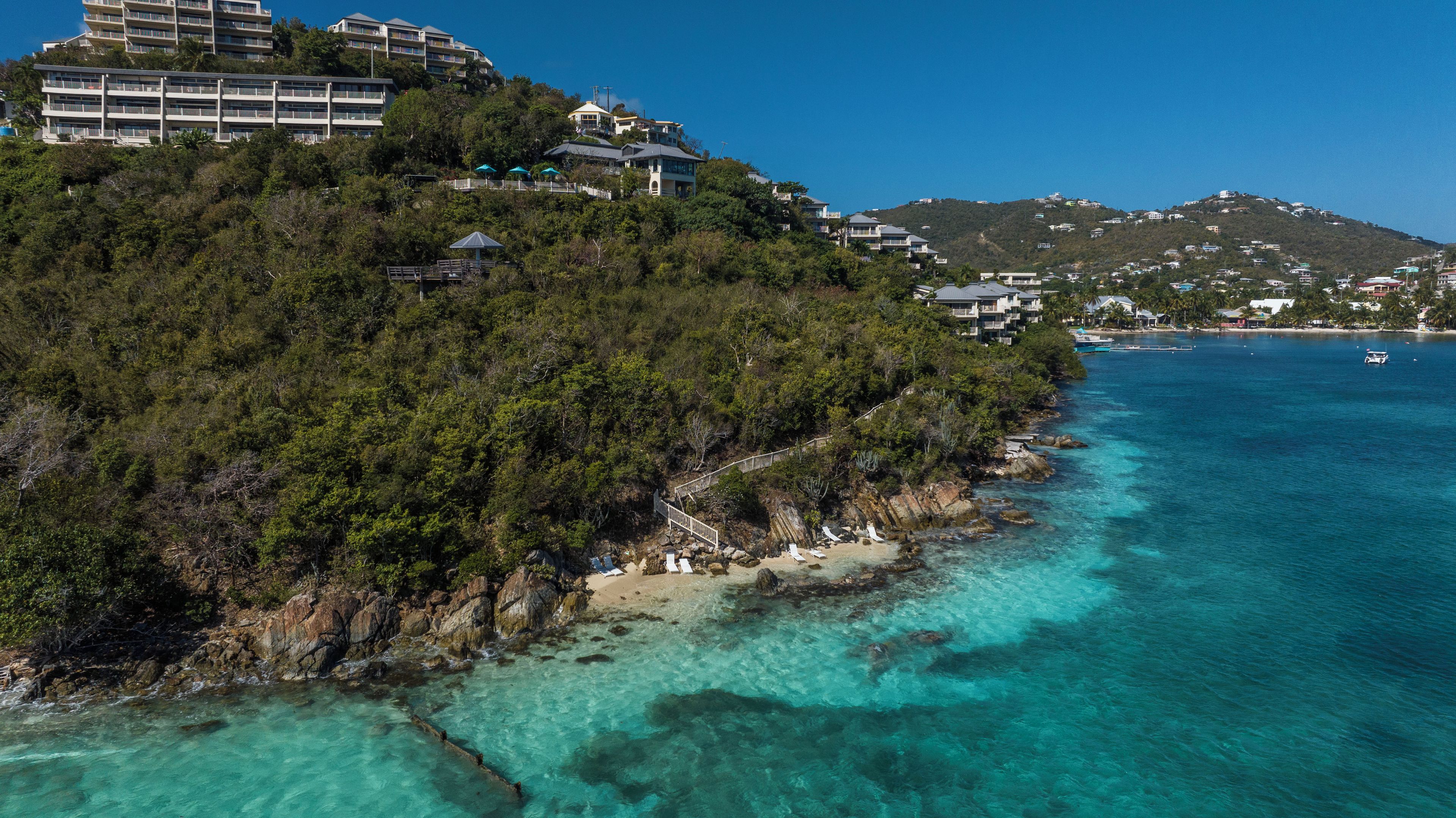 Une zone côtière avec une plage, des eaux turquoise et limpides, et une colline parsemée de bâtiments.