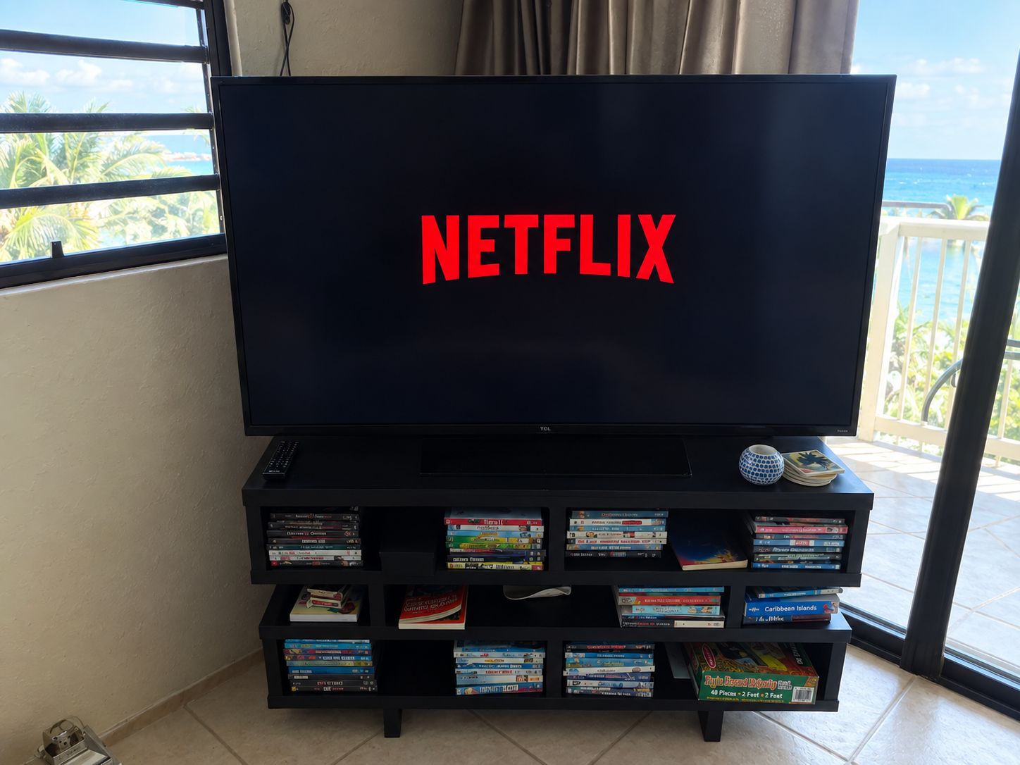 A television with the Netflix logo on the screen, placed on a black entertainment unit with a view of the ocean through a window.