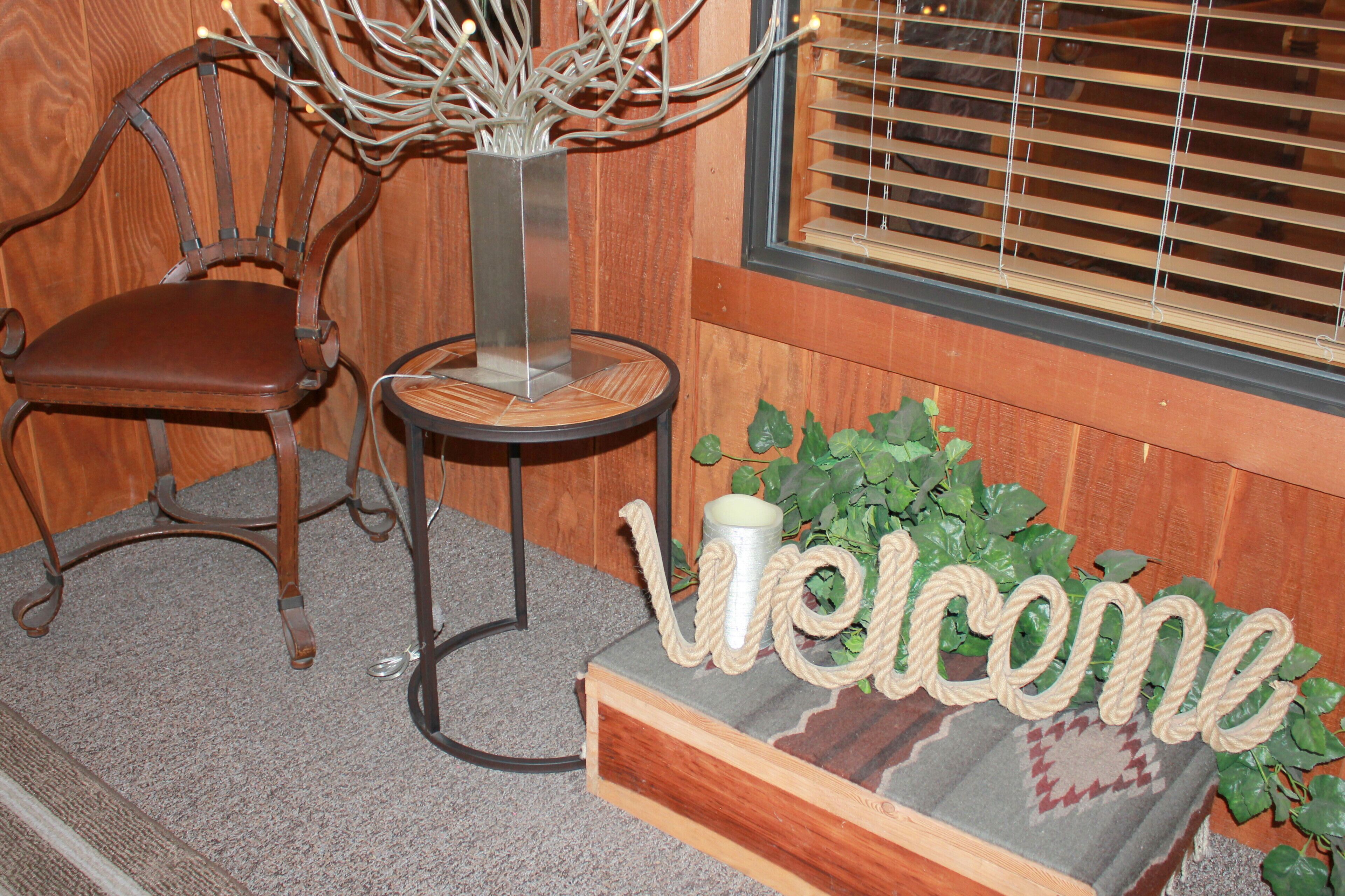 A wooden chair, a small table with a vase, and a welcome sign with rope letters.