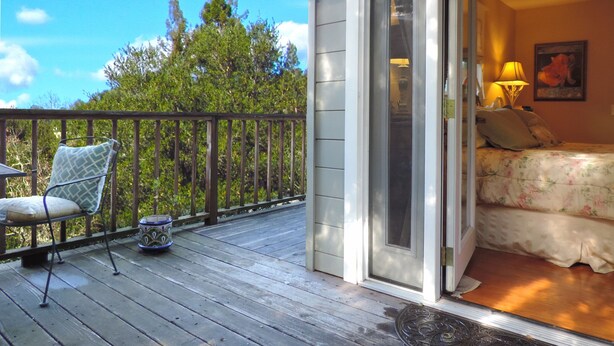 Un balcon avec une chaise et un pot décoratif, une porte-fenêtre coulissante donnant sur une chambre avec un lit et une lampe, et une vue sur les arbres et le ciel.