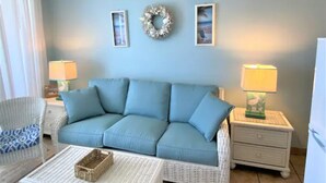 A living room with a blue sofa, two side tables with lamps, a wicker coffee table, a white chair with a blue cushion, and a wall with two framed pictures and a wreath. - Spectacular views, direct oceanfront, separate bedroom ! (Daytona Beach)