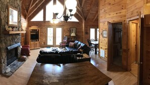 A cozy cabin interior with a stone fireplace, wooden walls, a dining area, and a large window. - Creek Front Mountain Cabin with Jacuzzi. (Andrews)