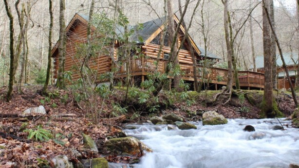 A log cabin with a wooden deck, surrounded by a forest and a flowing stream. - Creek Front Mountain Cabin with Jacuzzi. (Andrews)